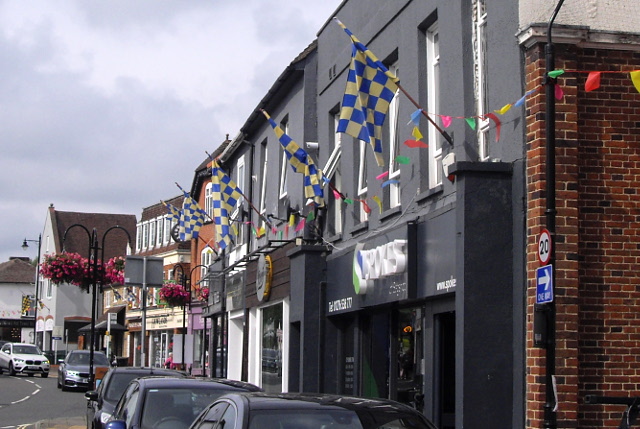 a grey painted shop front with checkered flags from its upper storey, and further shop fronts and hanging flower baskets visible further along.
